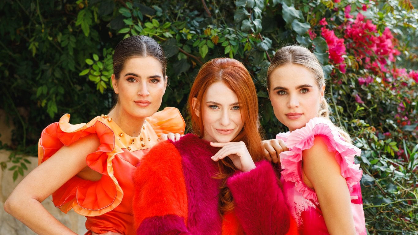 Haley J. debut collection — three models in a lush garden with bougainvillea, wearing the Cathy Orange Pleated Mini Dress, Tina Faux Fur Coat in red and pink, and Pink Pleated Party Top with Pink Pleated Party Pant