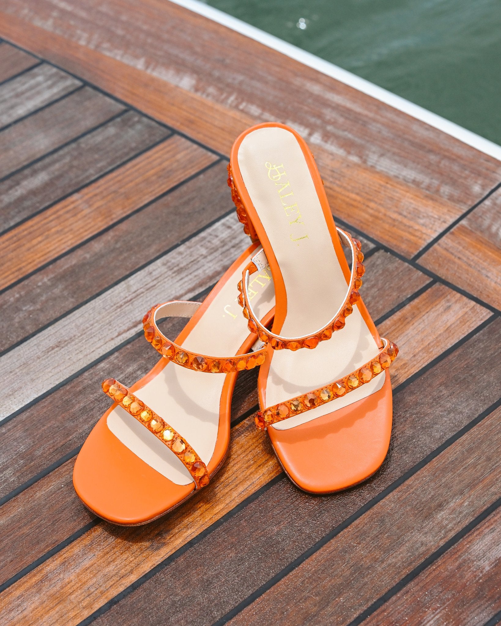 Haley J. Katie Orange Crystal Heel — orange open-toe double-strap mule sandals with amber crystal trim along the straps and low block heel, displayed on a wooden dock with water in the background