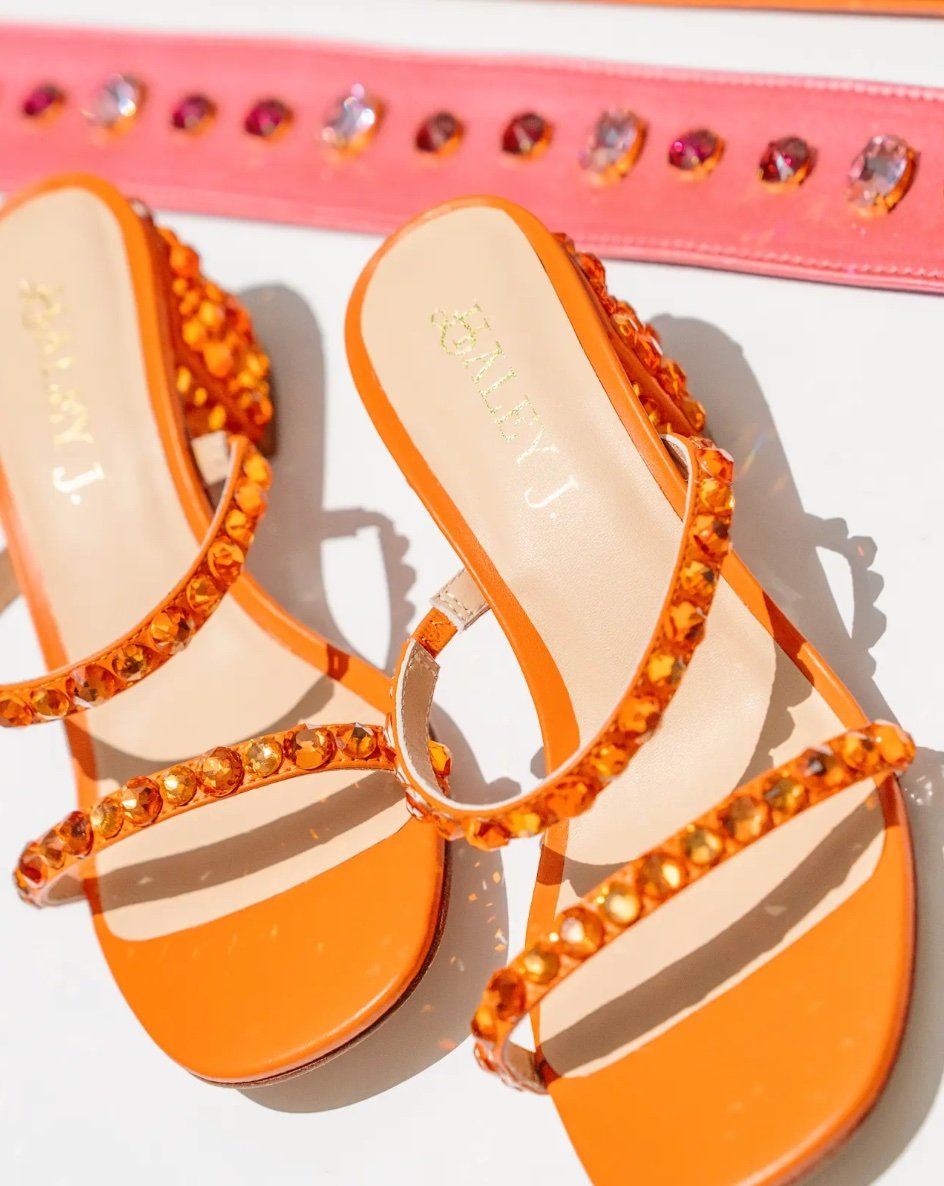 Haley J. Katie Orange Crystal Heel — flat lay of orange double-strap sandals lined with amber crystal embellishments, Haley J. branded insole, styled alongside a pink crystal-studded party belt