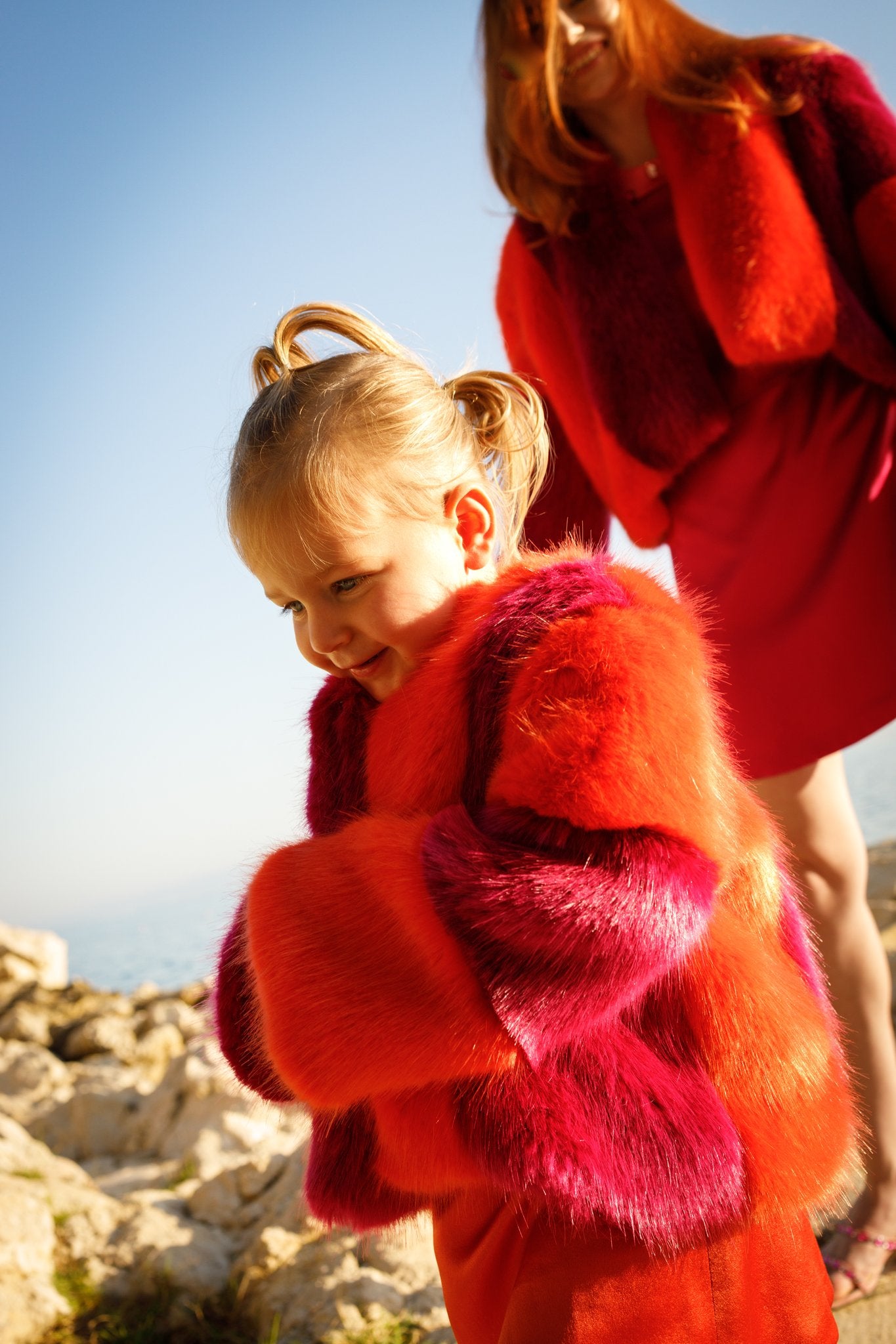 Haley J. Mini Tina Faux Fur Coat — girl's two-tone red and hot pink faux fur coat, worn with a red outfit, smiling on coastal rocks with a woman in a matching adult Tina Faux Fur Coat in the background