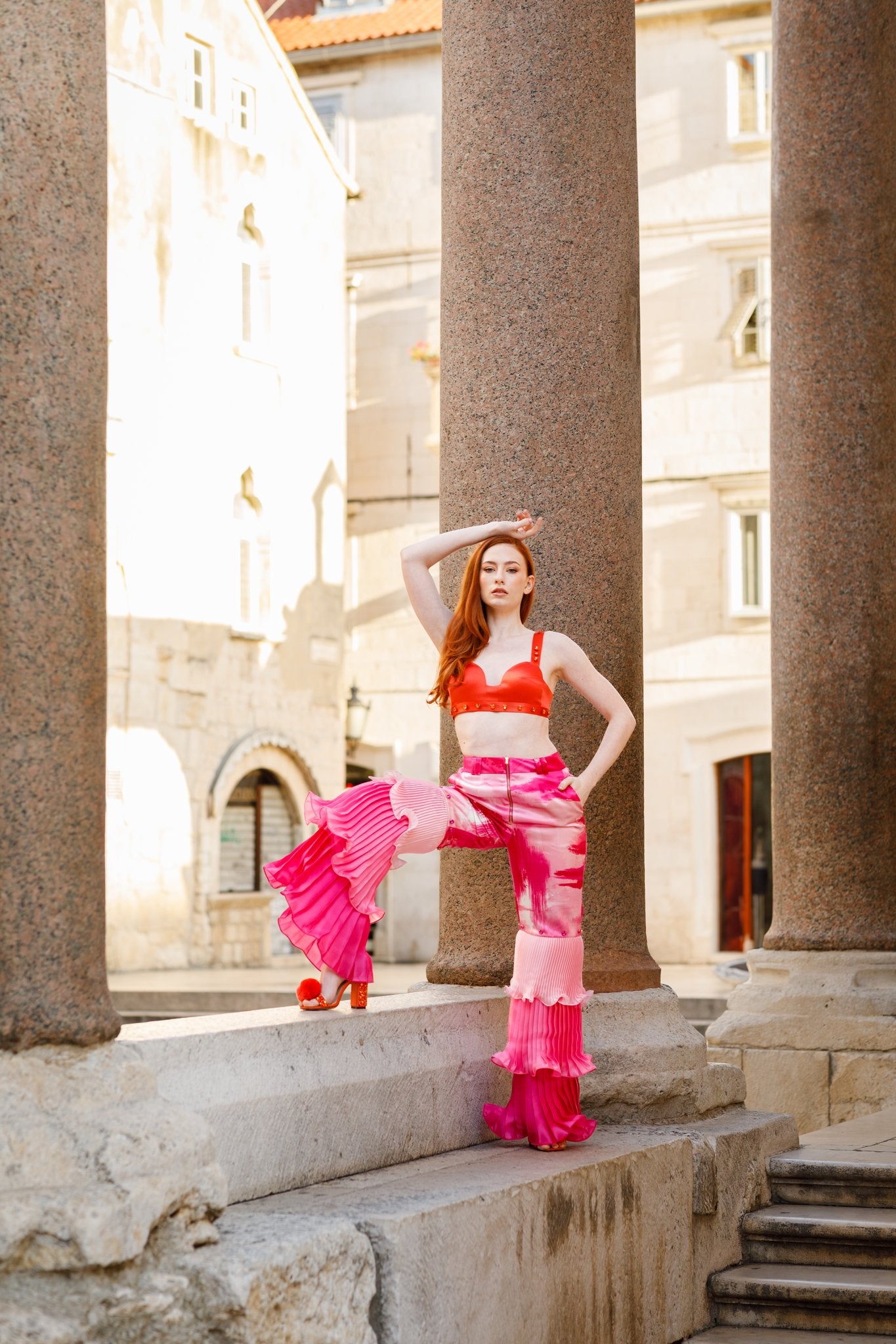 Haley J. Pink Pleated Party Pant styled with her orange studded bra top — abstract pink print wide-leg trousers with dramatic pleated ruffle hem, paired with orange pom pom heels among stone columns of a European plaza