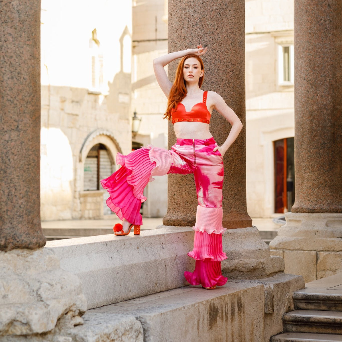 Haley J. Pink Pleated Party Pant styled with her orange studded bra top — abstract pink print wide-leg trousers with dramatic pleated ruffle hem, paired with orange pom pom heels among stone columns of a European plaza