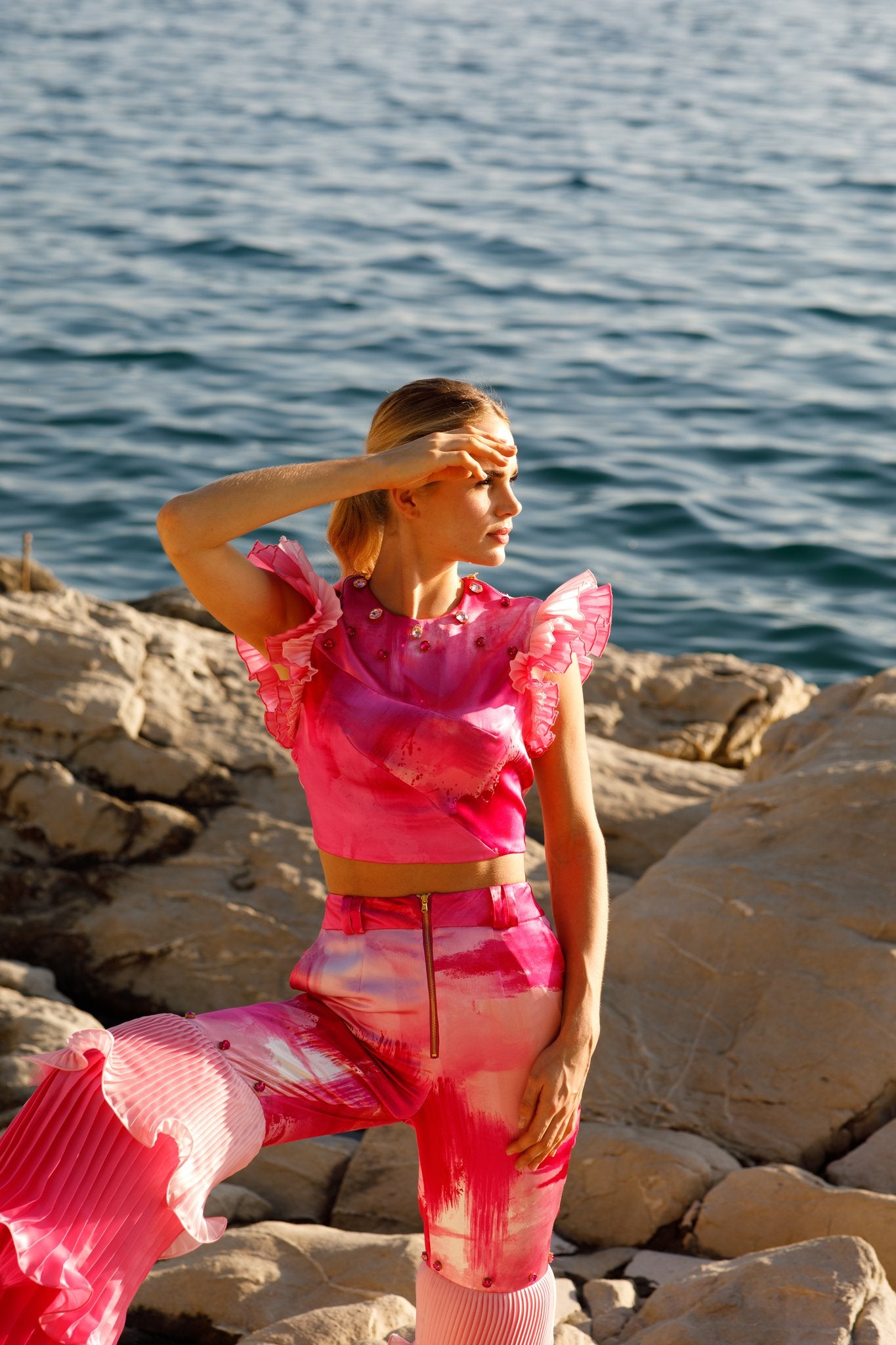 Haley J. Pink Pleated Party Top and Pink Pleated Party Pant — abstract pink print co-ord set featuring a ruffle-sleeve crop top with crystal embellishments and wide-leg pleated trousers with gold zip detail, styled on coastal rocks overlooking the sea