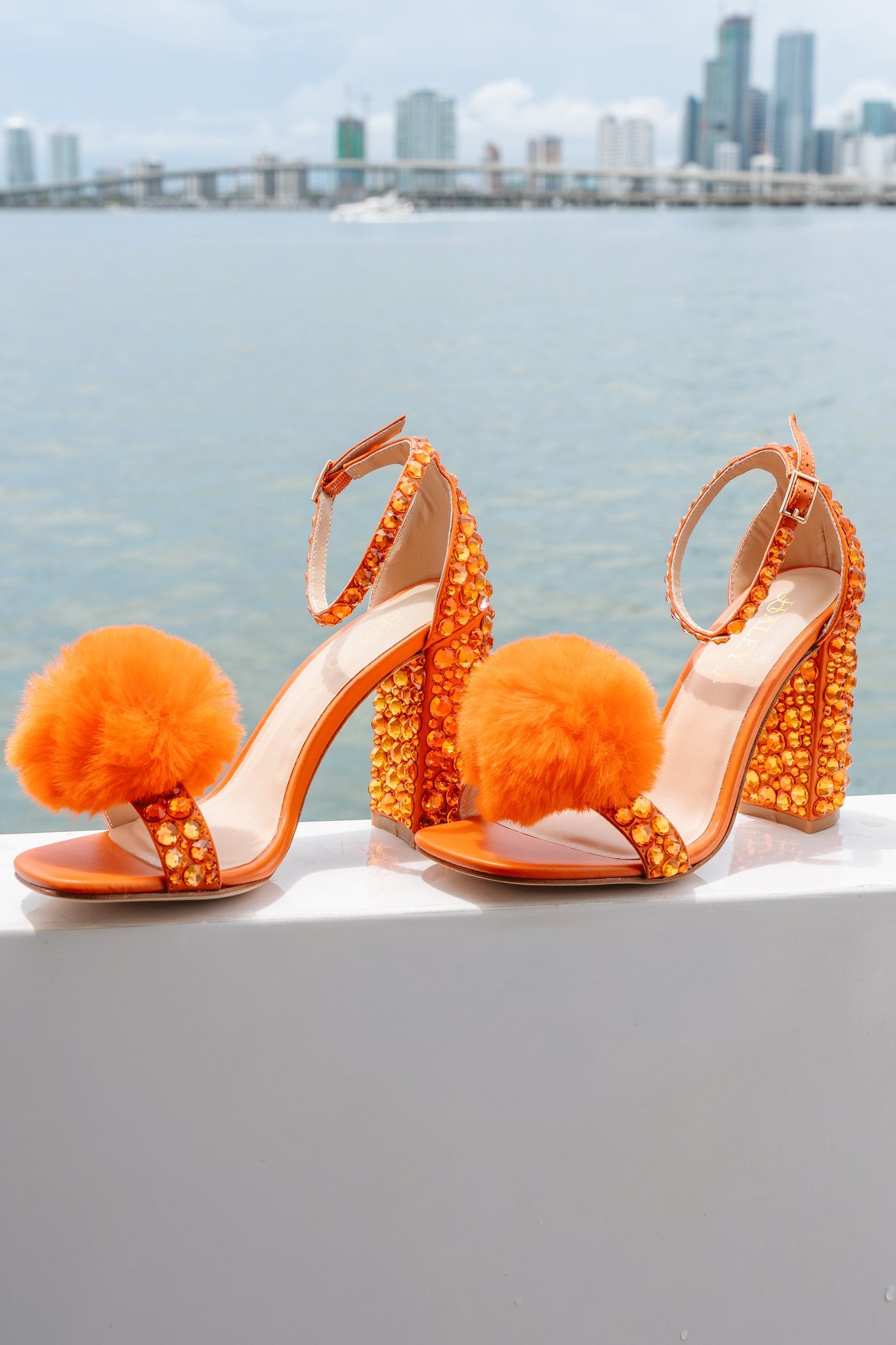 Haley J. Pom Pom Orange Party Shoe — bold orange block heels covered in amber crystals with fluffy orange pom pom toe strap and ankle buckle detail, displayed against a Miami waterfront skyline