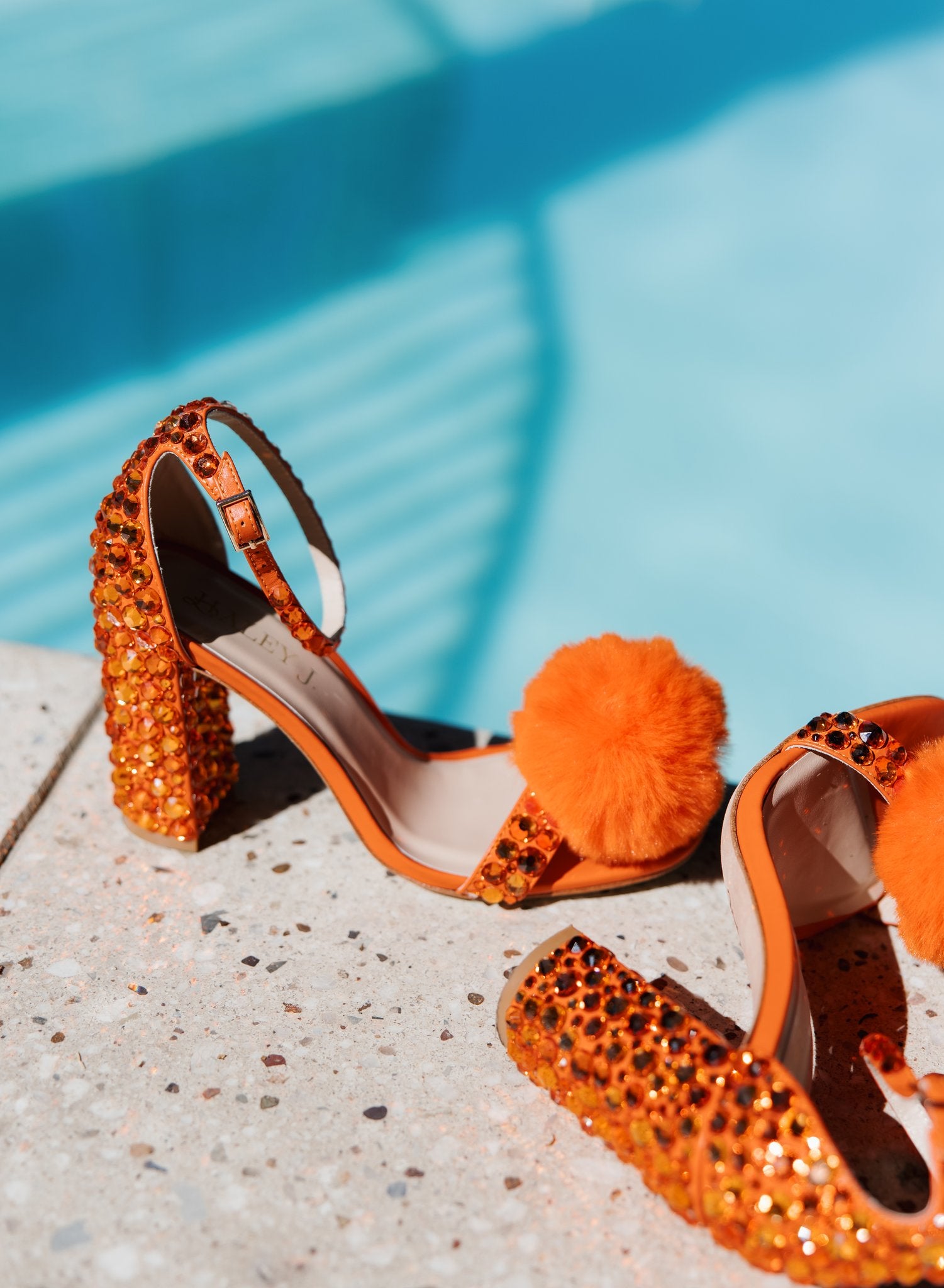 Haley J. Pom Pom Orange Party Shoe — close-up of vibrant orange ankle strap heels fully encrusted in orange crystals with fluffy orange pom pom toe detail, displayed beside a sunlit pool