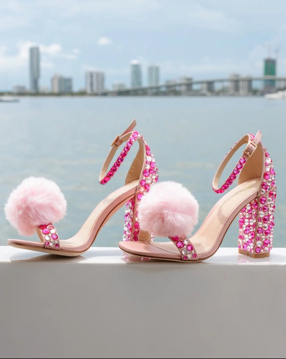 Haley J. Pom Pom Pink Party Shoe — blush pink ankle strap heels with hot pink and clear crystal embellishments and fluffy pink pom pom toe detail, displayed against a Miami waterfront skyline