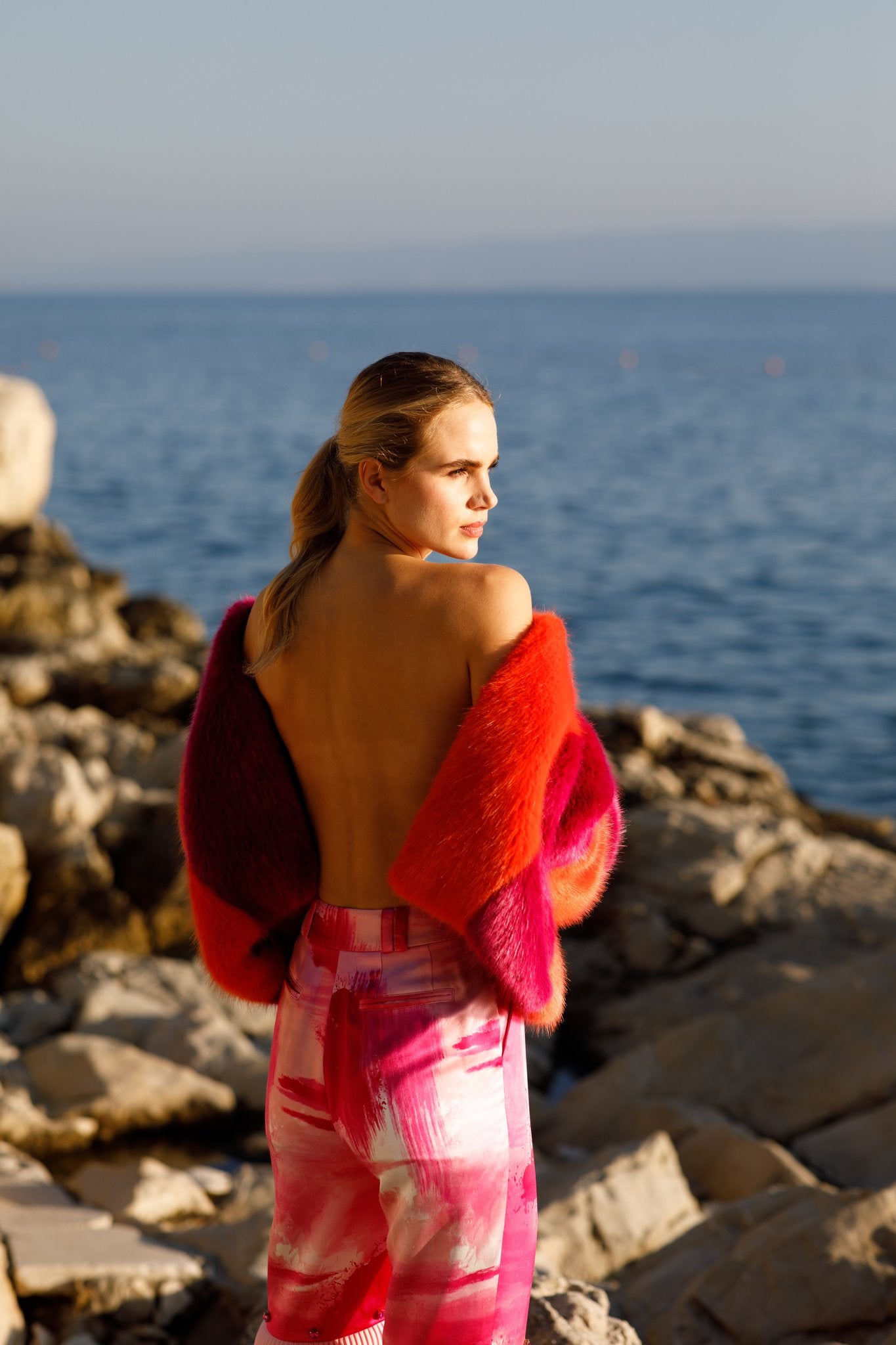 Haley J. Tina Faux Fur Coat — two-tone orange and hot pink faux fur coat worn off-shoulder, paired with abstract pink print trousers, back view against a coastal rock and ocean backdrop