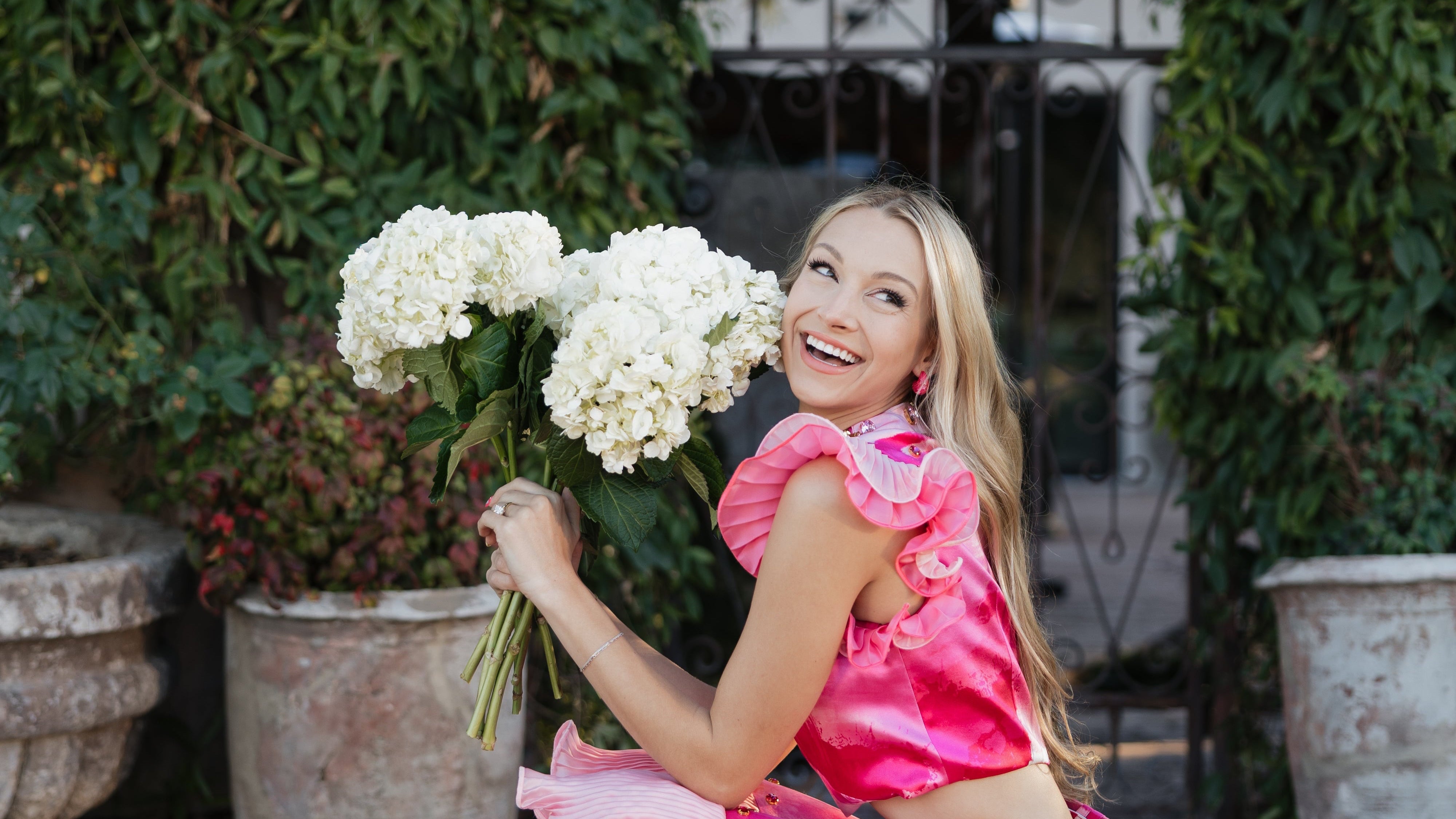 Woman in a pink dress holding flowers in an outdoor setting with greenery.